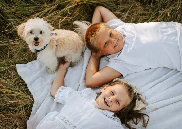 A shot of a young boy and girl lying on a white blanket in tall grass alongside their dog.